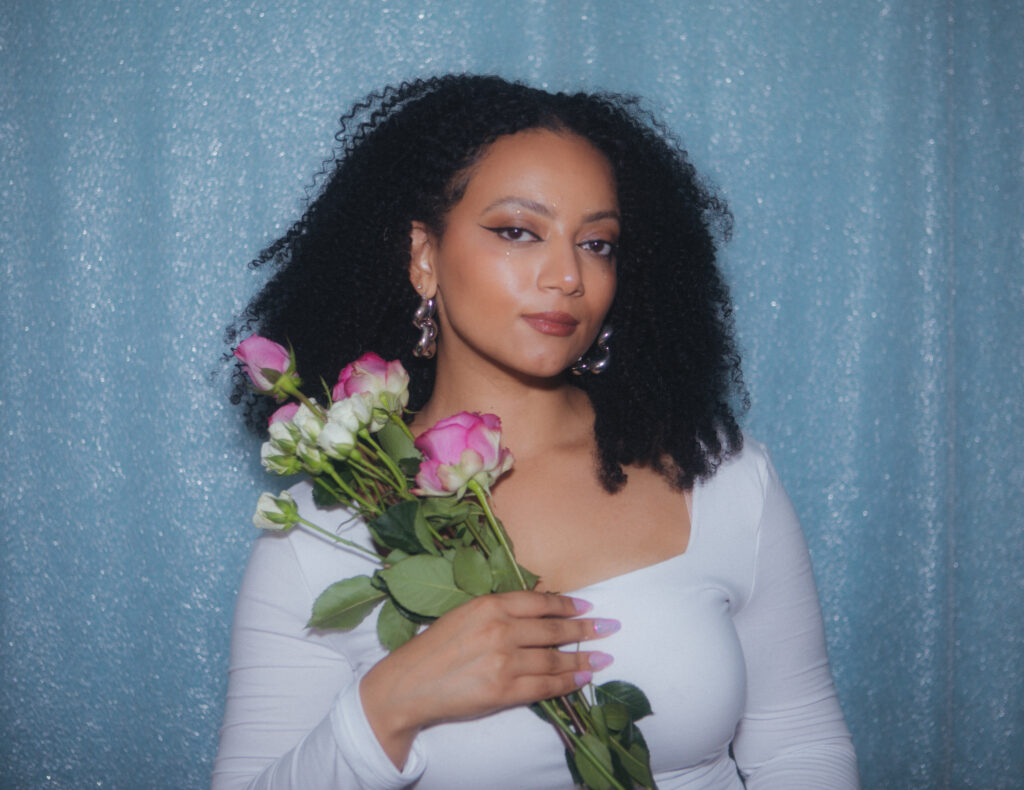 Author Meghan Malachi looking lovely with her hair flowing holding a bunch of roses and wearing a white top. She has light brown skin and dangly earrings, and big lovely eyes.