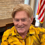 Author R. J. Lee wearing a bright yellow shirt. He's a handsome man with strawberry blond hair, a strong jaw and an impish smile.