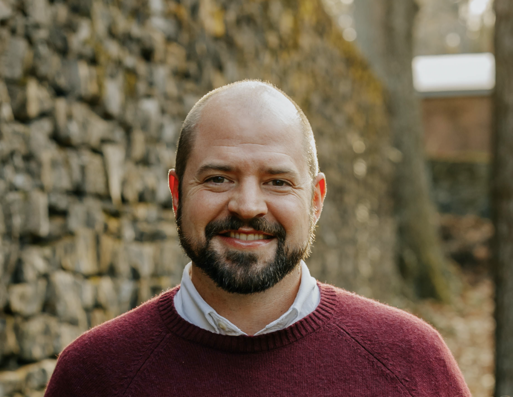 Author Noah Soltau wears a red sweater. He smiles with a close-trimmed beard. His head is bald, and he stands beside a brick wall.