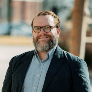 Author Derek Prall. He has red hair, combed back with a salt and pepper beard and glasses. He wears an open-collared blue shirt under a navy-blue blazer. The background is blurry.