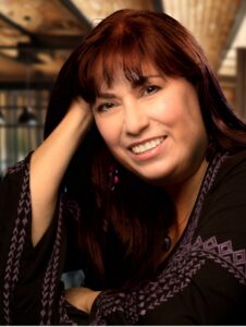 Author Suzanne Mattaboni smile with her head resting on her hand. She has long dark hair with bangs and an open welcoming smile.