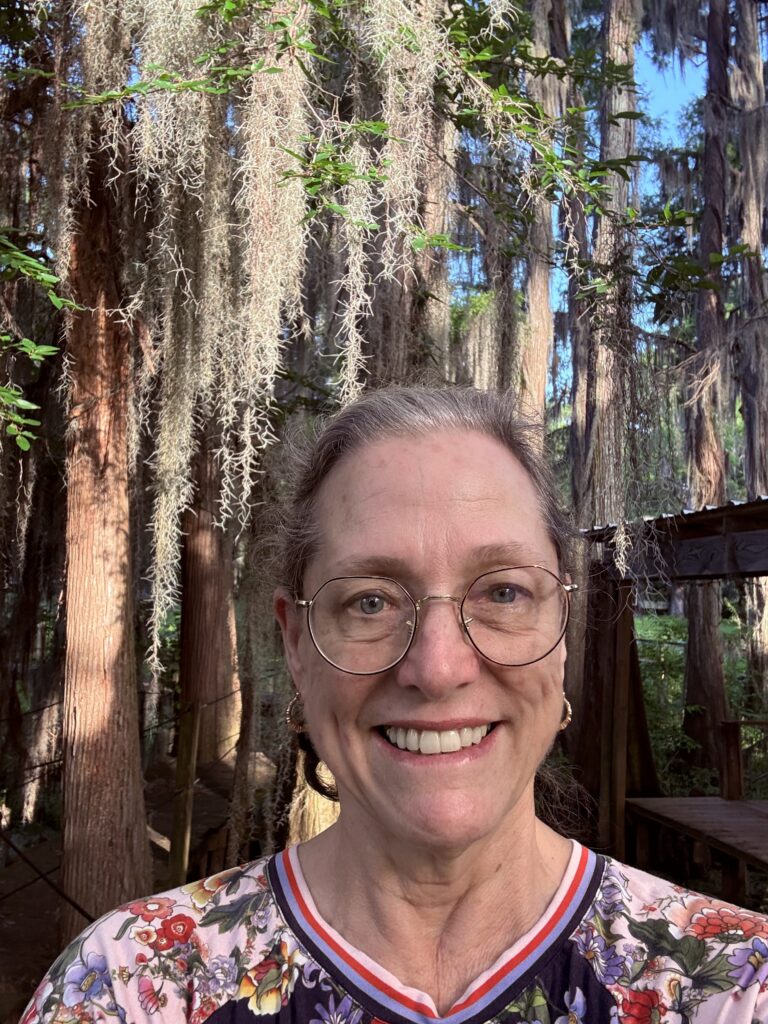 Kim Davis. Smiling woman in flowered t-shirt with Cypress trees in background. She wears wire framed glasses with brown/gray hair pulled back.