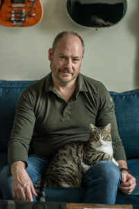 Author Sean Carberry sits smiling at the camera wearing a long-sleeved army-green t-shirt and holding a tabby cat on his lap.