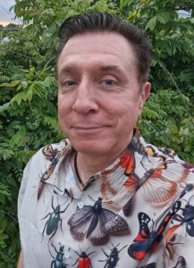 David B. Prather, 2026 Arthur Smith Poetry Prize Winner. He is a dark haired, middle-aged white man with a cheeky grin, wearing a shirt with a pattern of brightly-colored insects. 