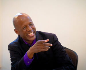 Author Steven T. Moore smiling and pointing at an audience member at one of his poetry readings at the TACW conference in Granbury, Texas, 2026. He is a man with a shaved head, dark brown skin, and he wears a black suit. His smile is disarming.