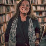 Author, editor, publisher Kimberly Parish (K.P.) Davis. Here she is in a room filled with books, wearing wire framed glasses and grinning. Her haire is long and unkempt.