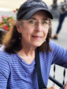Elaine Fowler Palencia, poet and author. She wears a ball cap and a blue striped shirt. He has shoulder-length brown hair and wire framed glasses, and a hesitant but welcoming smile--like she is in the middle of a conversation. Blurry background shows people in a city-setting.