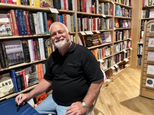 Author Michael Simms sitting and smiling in a bookkstore. He has a white beard and no hair on top.