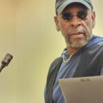 Author Earl S. Braggs at the podium turns to look at the camera. He is wearing a ball cap and dark glasses. 
